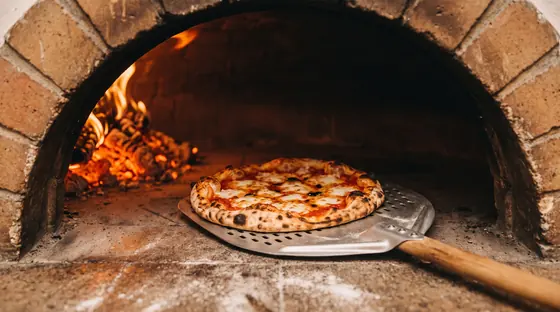 Ein gelochter Pizzaschieber holt eine Pizza aus dem gemauerten Holzofen, hinten Glut und Flammen